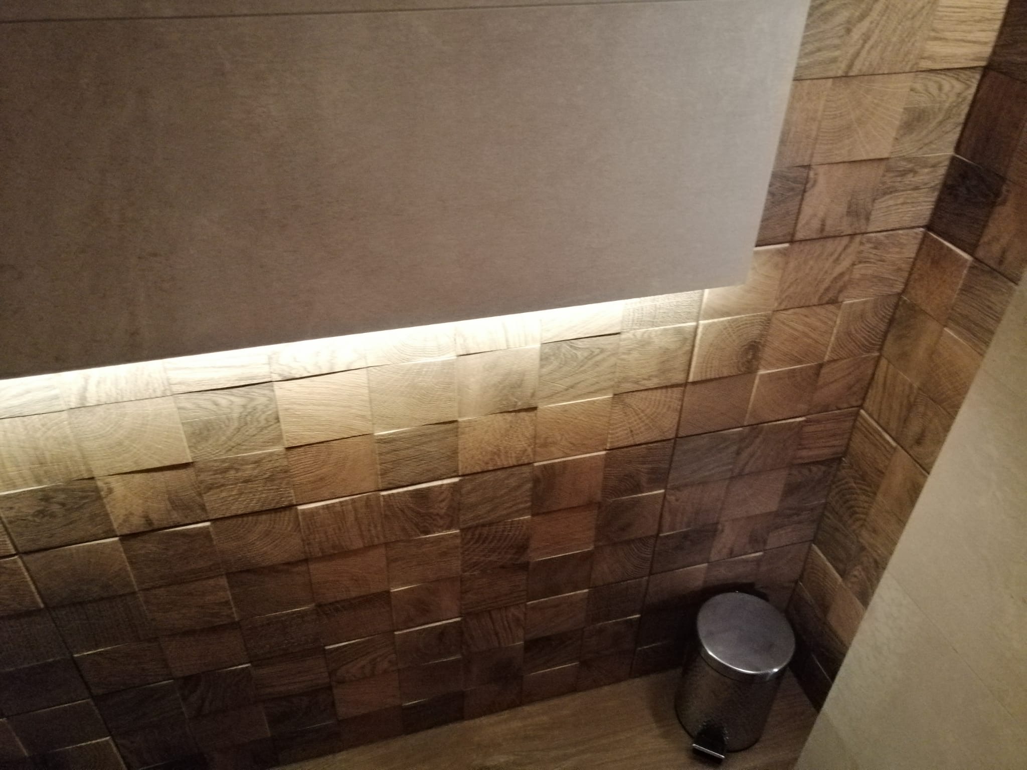 Ściana łazienki wyłożona drewnianymi, kwadratowymi płytkami z widocznym usłojeniem, podświetlona od góry listwą LED, z metalowym koszem na śmieci w rogu.