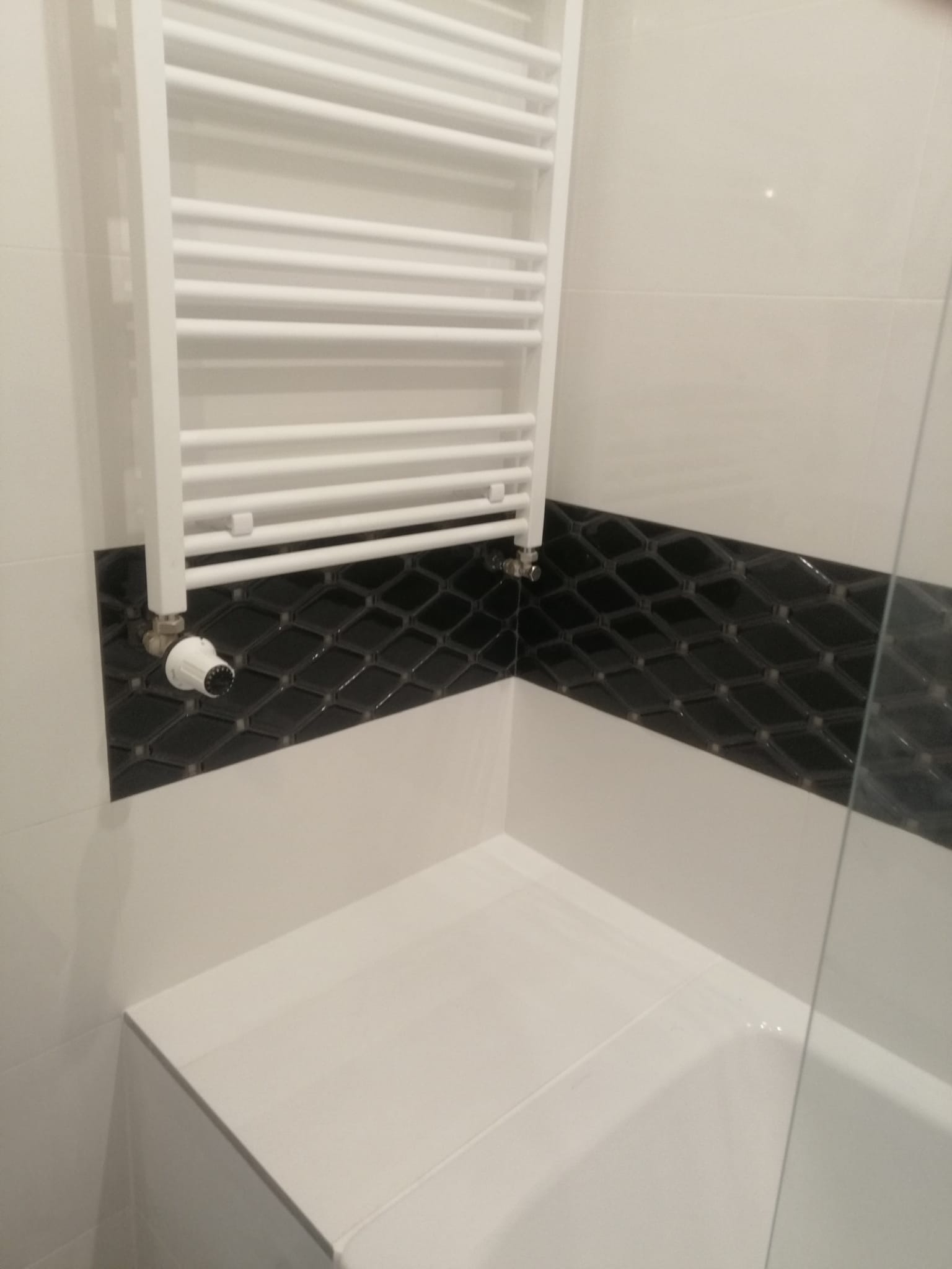 Fragment wykończonej łazienki z białymi i czarnymi płytkami, grzejnikiem drabinkowym oraz widoczną krawędzią wanny za szybą.