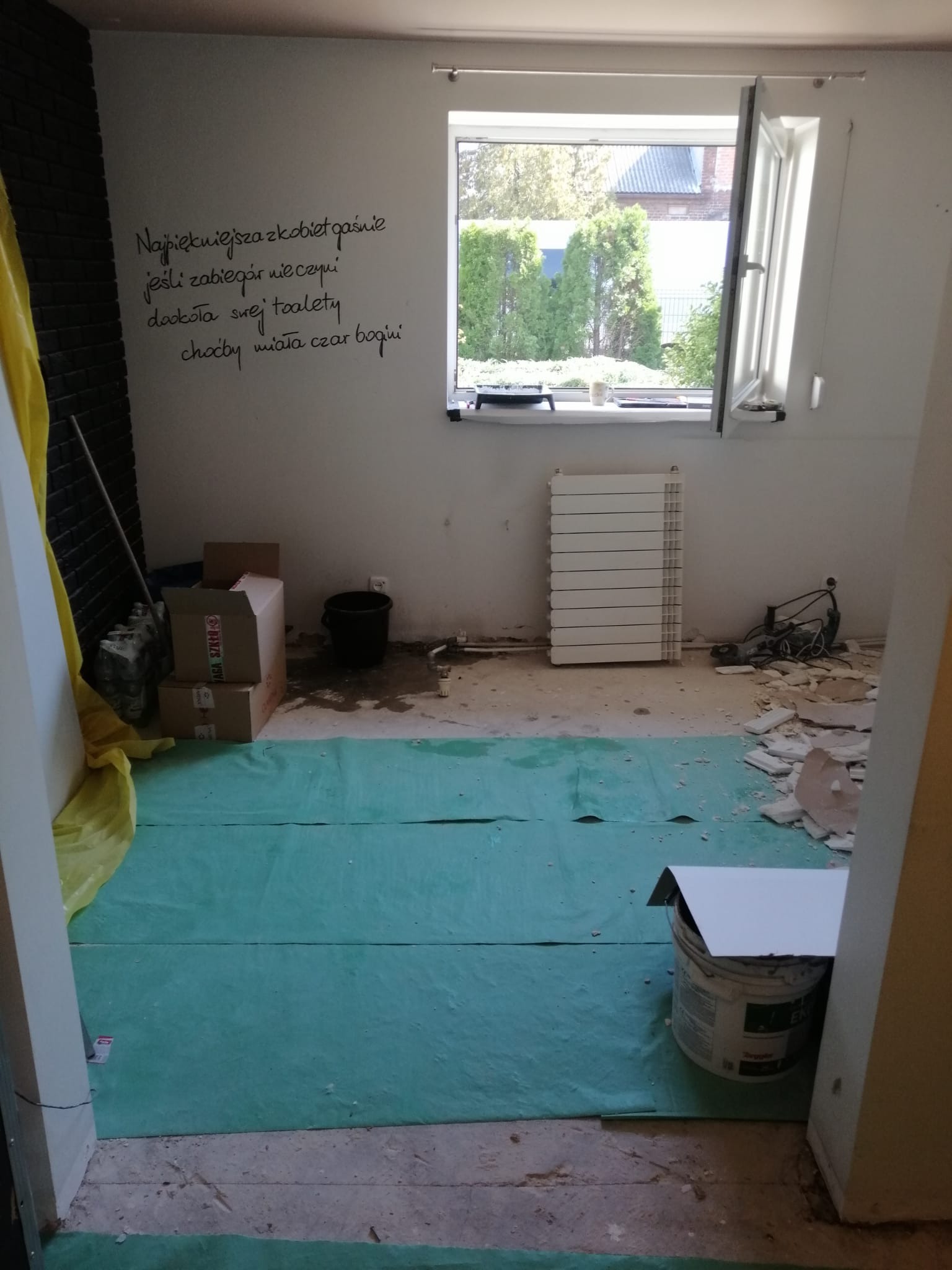 Remontowane pomieszczenie z zieloną folią ochronną na podłodze, widoczny grzejnik, okno z widokiem na zieleń i fragment ściany z napisanym czarnym markerem wierszem.