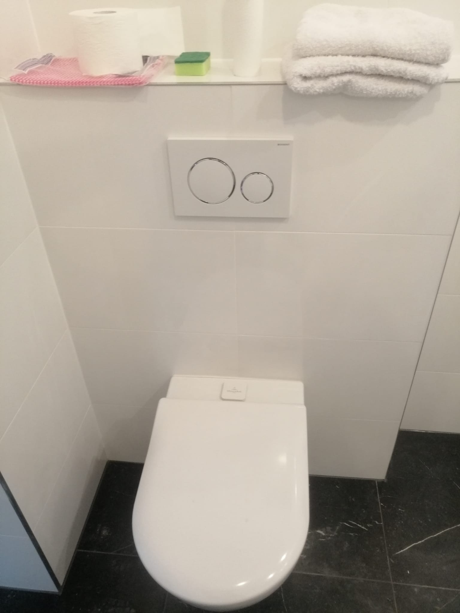 Biała toaleta podwieszana z przyciskiem spłukującym na białych płytkach, nad nią półka z ręcznikami, papierem toaletowym i gąbką, czarna marmurkowa podłoga.