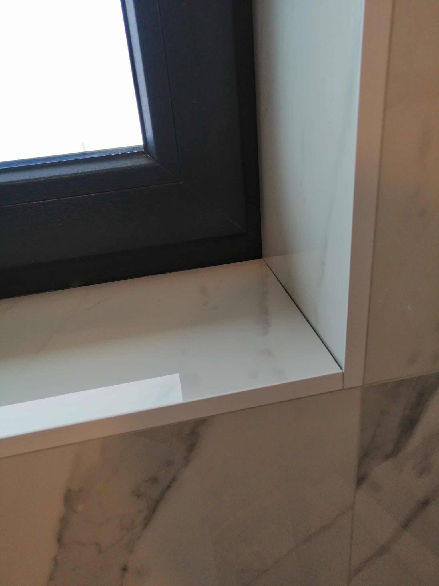 Fragment okna z czarną ramą i marmurowym parapetem, wykończonymi płytkami imitującymi marmur na ścianie.