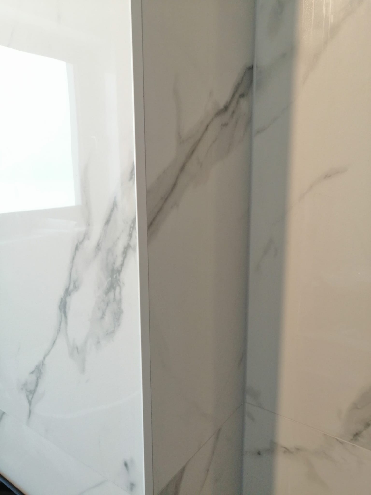 Fragment łazienki wykończonej białymi płytkami imitującymi marmur z wyraźnymi szarymi żyłami, widoczne narożniki i fuga.