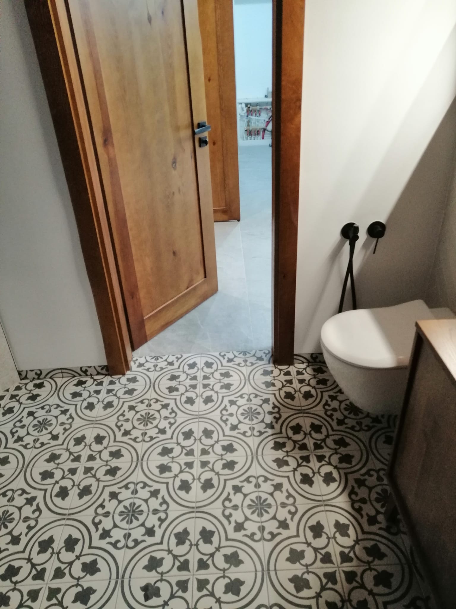 Ukończona łazienka z wzorzystą, czarno-białą podłogą, drewnianymi drzwiami z czarnymi klamkami i toaletą z czarną armaturą.