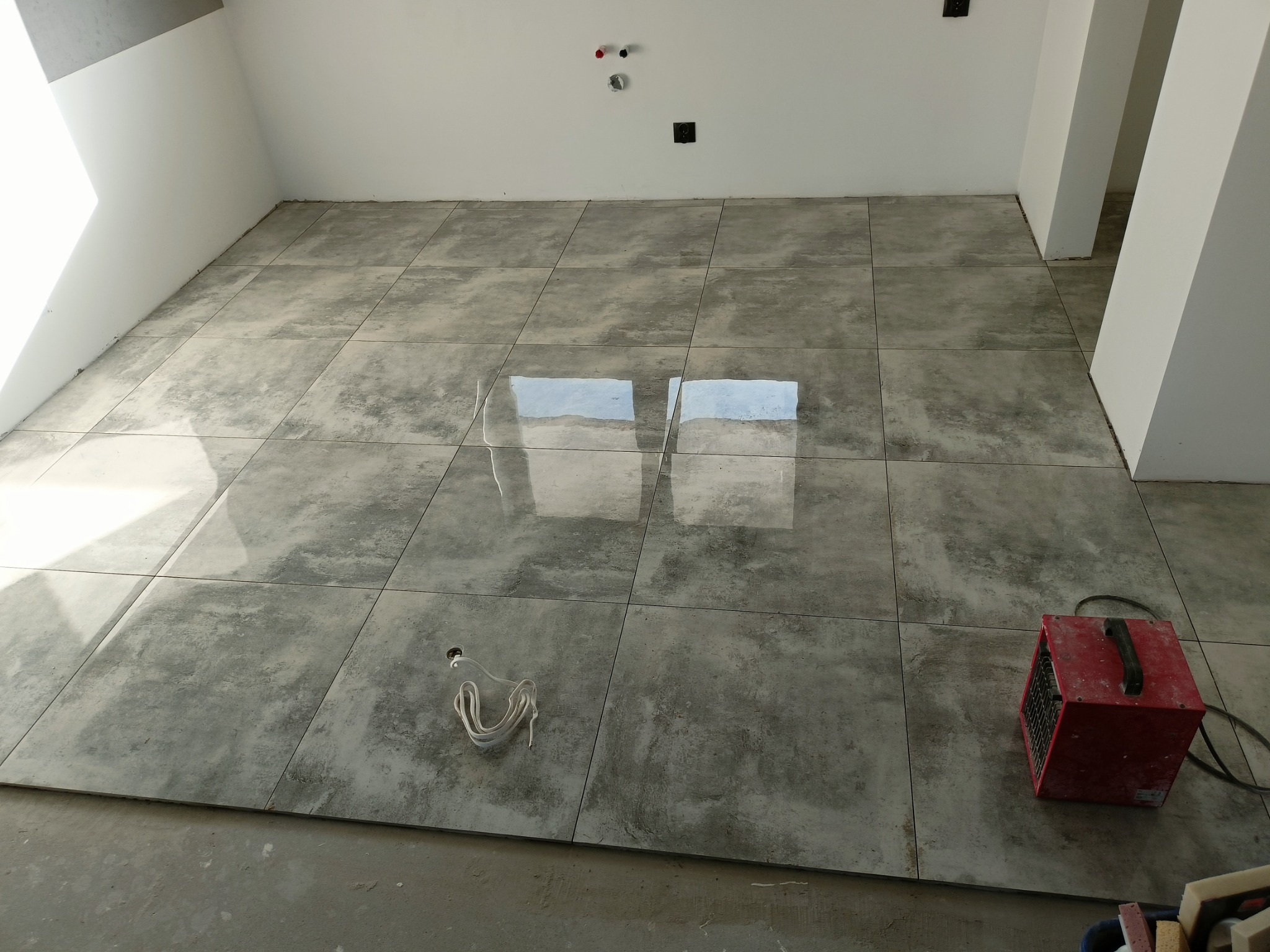 Świeżo ułożone, duże, szare płytki podłogowe w pustym pomieszczeniu, z widocznym grzejnikiem elektrycznym i wystającym przewodem. Na ścianie widoczne puszki elektryczne.