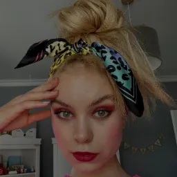 Portret młodej kobiety z blond włosami upiętymi w kok, w różowym swetrze i z kolorową apaszką na głowie. Wyrazisty makijaż oczu i ust. Ujęcie z bliska.