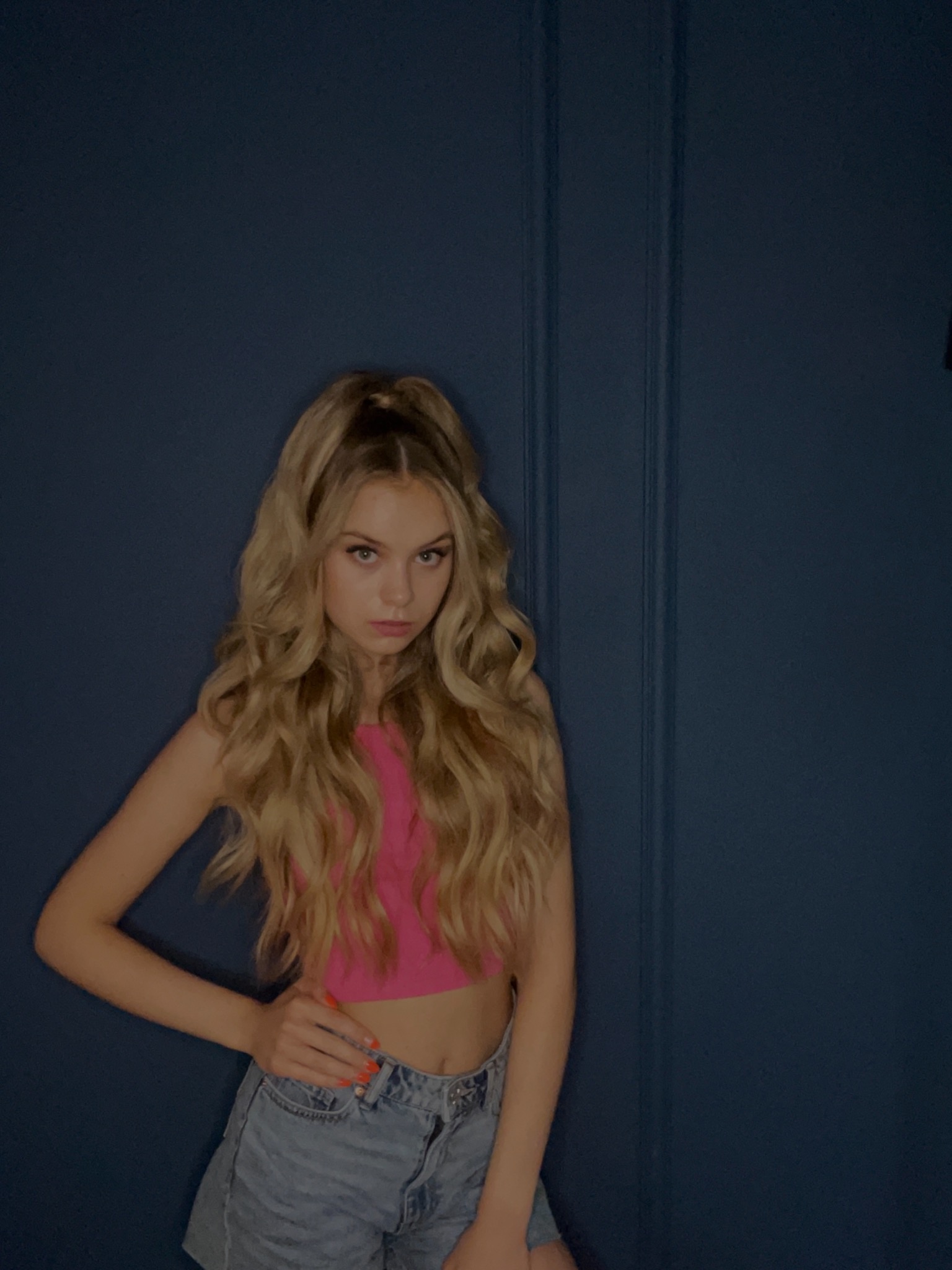 Portret młodej kobiety z blond włosami, ubrana w różowy top i dżinsowe szorty, pozująca na tle ciemnoniebieskiej ściany z pionowymi panelami.