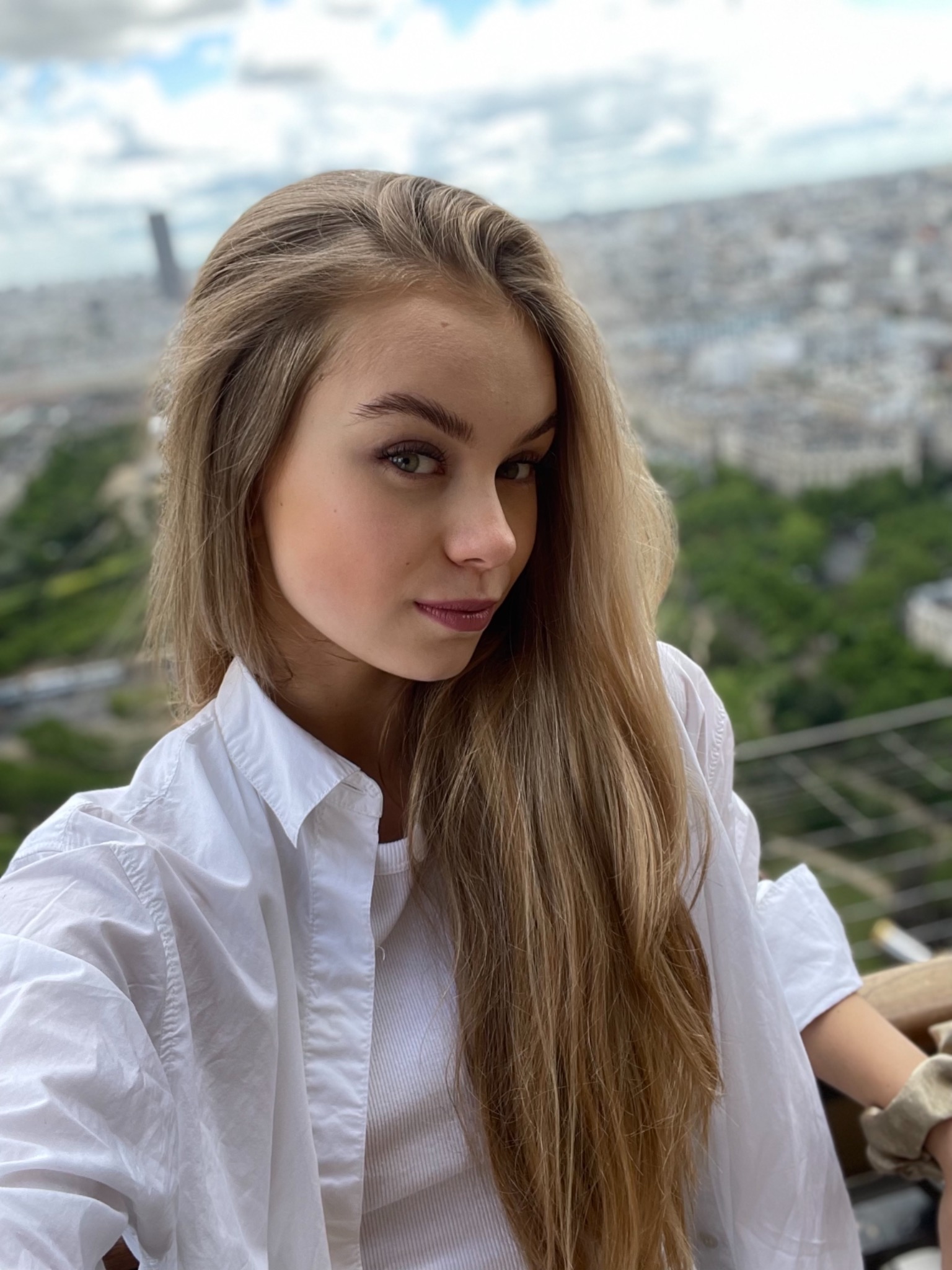 Portret młodej kobiety w białej koszuli, z widokiem na panoramę miasta w tle, uchwycony z lekkiego ukosa.