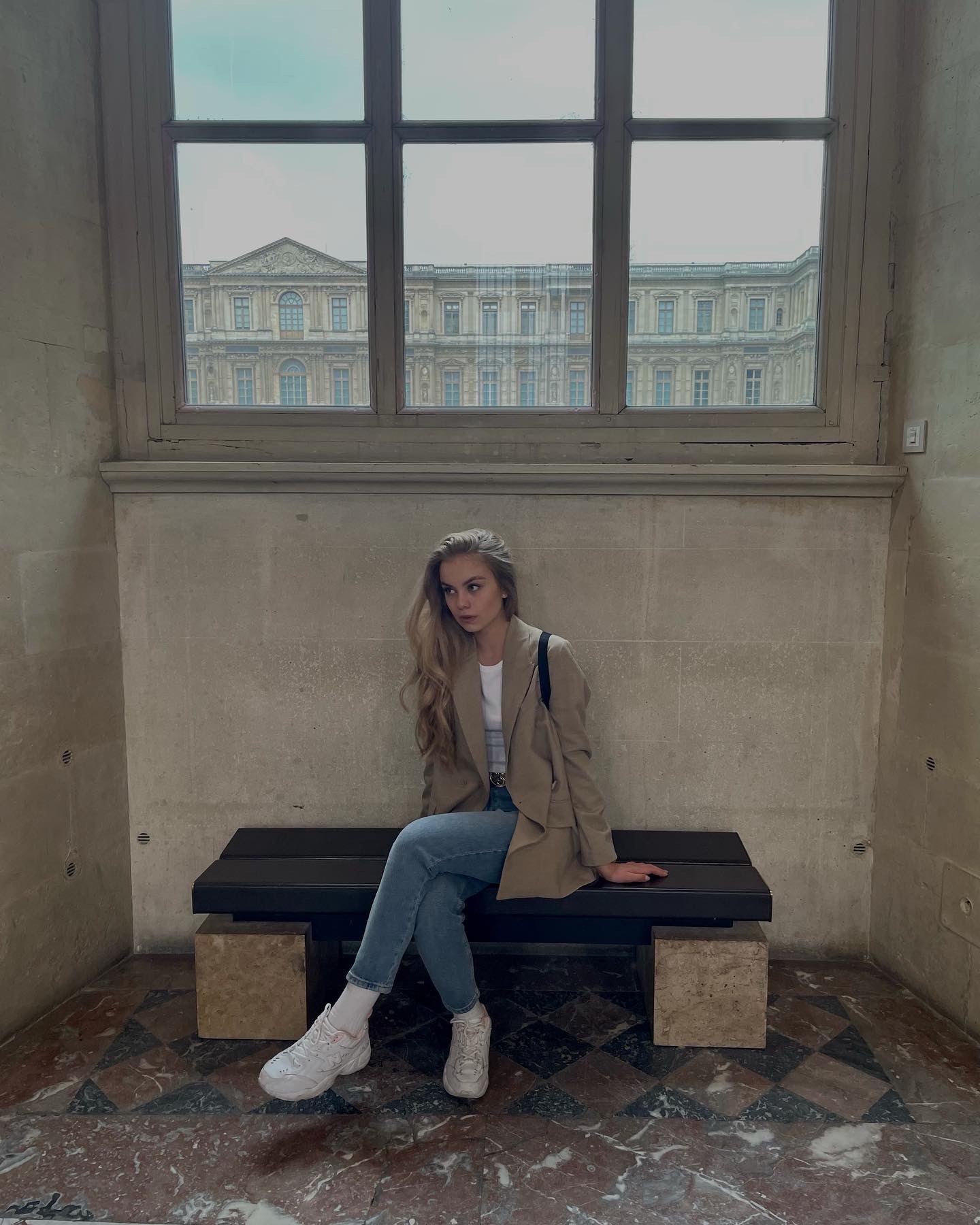 Kobieta w beżowej marynarce i jeansach siedzi na ławce na tle okna z widokiem na budynek w stylu klasycystycznym.