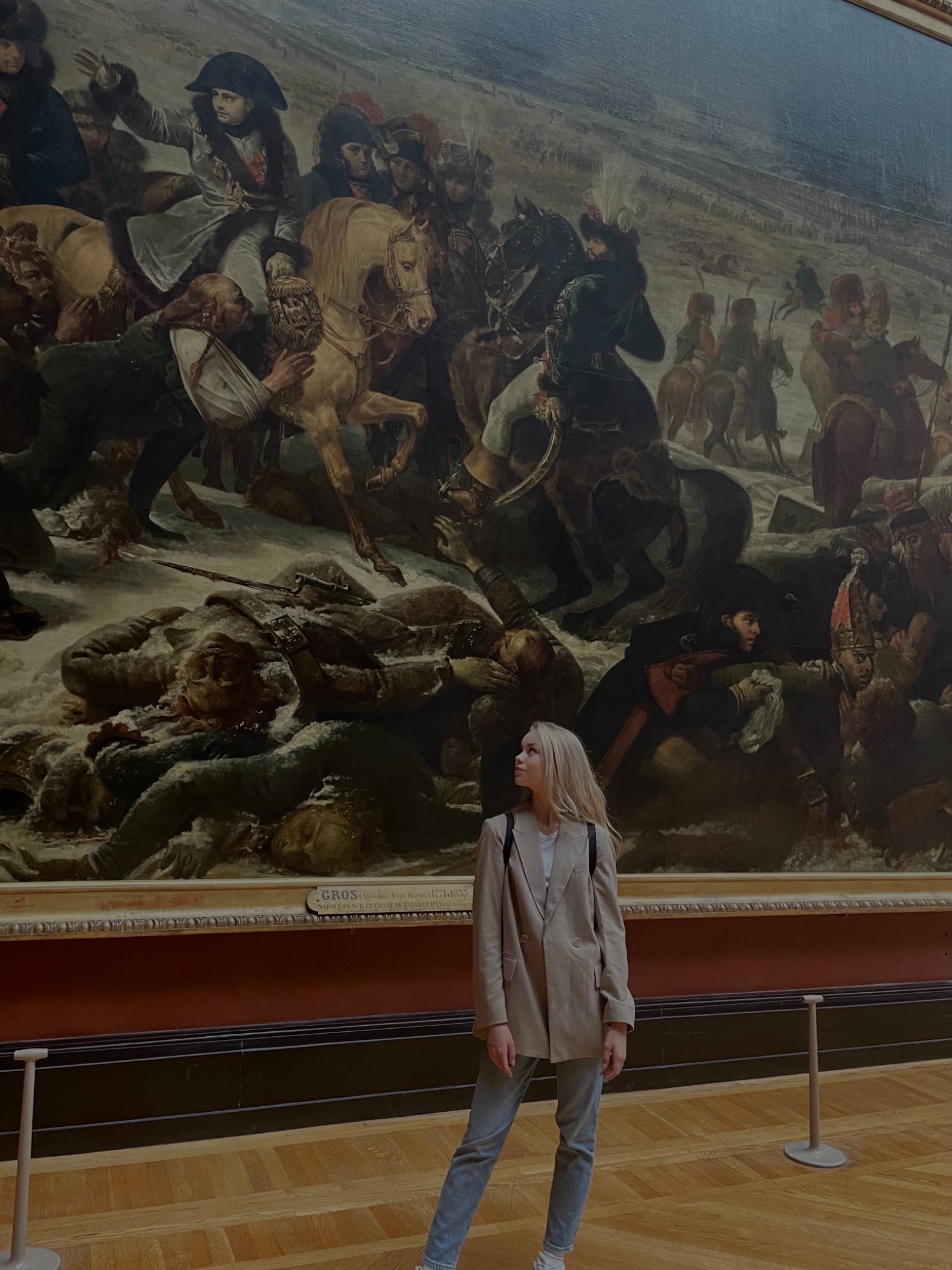 Kobieta w beżowej marynarce i jeansach stoi na tle ogromnego obrazu przedstawiającego bitwę z Napoleonem na koniu, w muzeum z drewnianą podłogą.