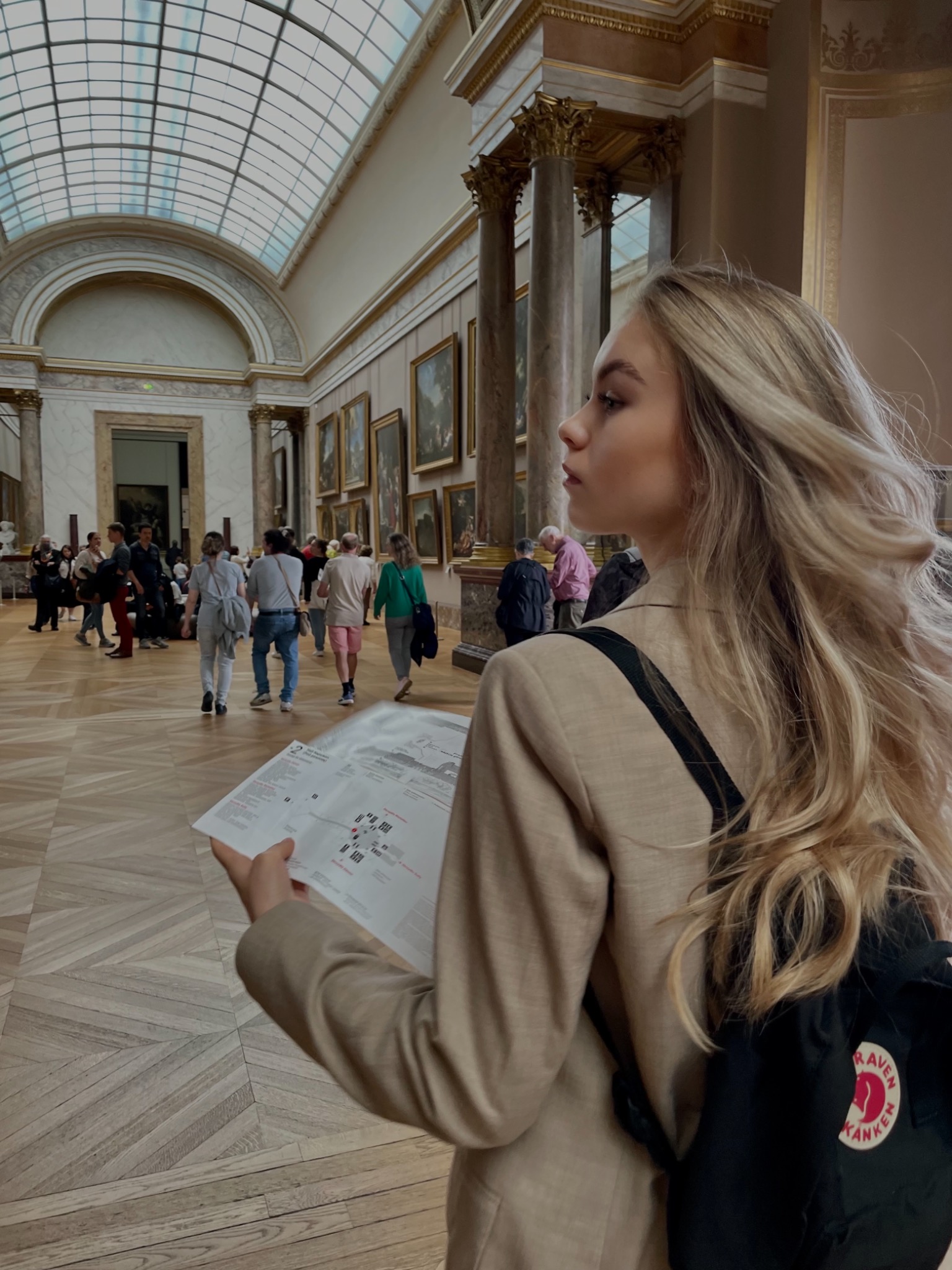 Portret kobiety z blond włosami w muzeum, trzymającej mapę, z kolumnami i obrazami w tle.