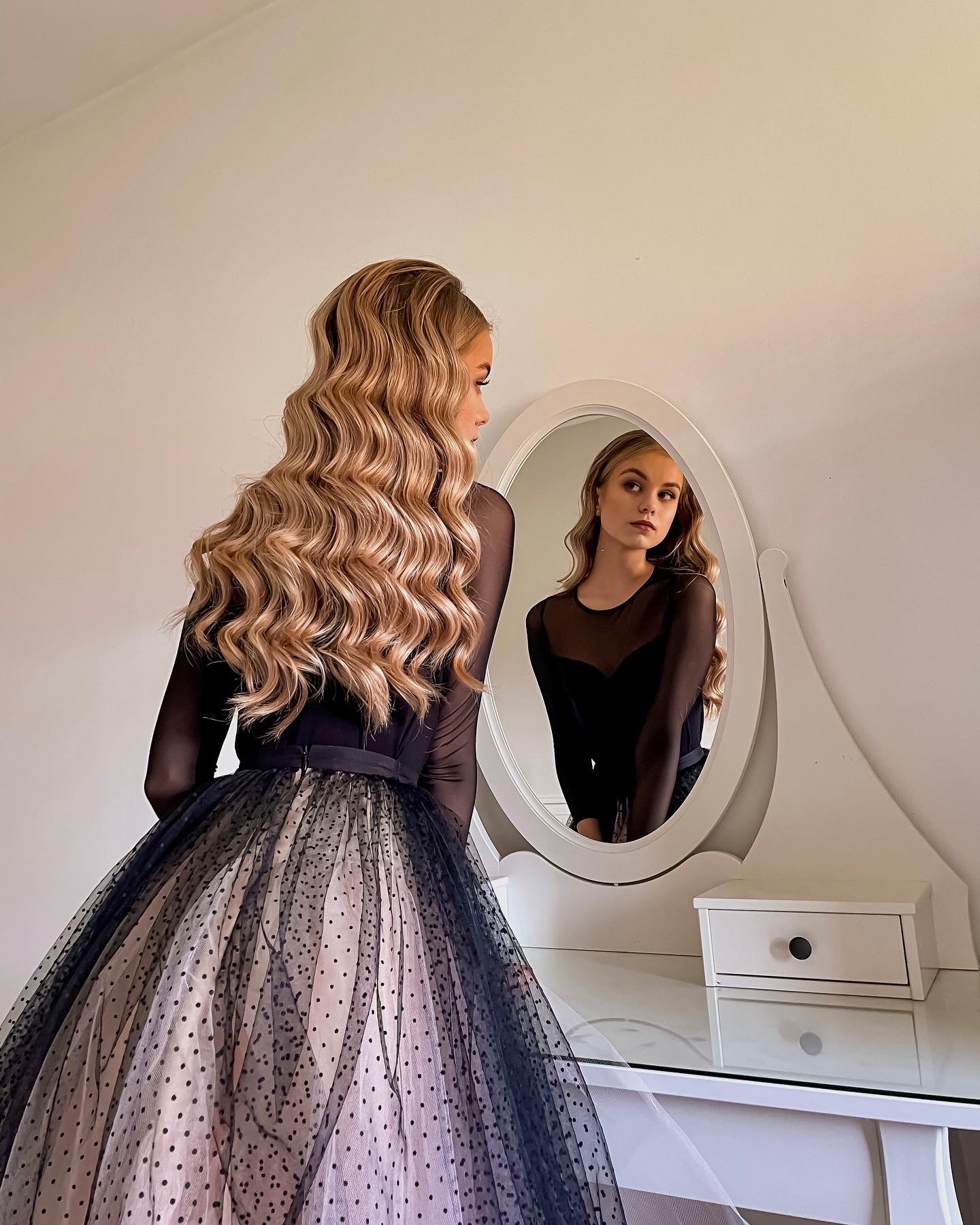 Kobieta w eleganckiej sukni z tiulowym dołem w kropki, stojąca przed toaletką z owalnym lustrem i patrząca w swoje odbicie, z widocznymi blond, falowanymi włosami.