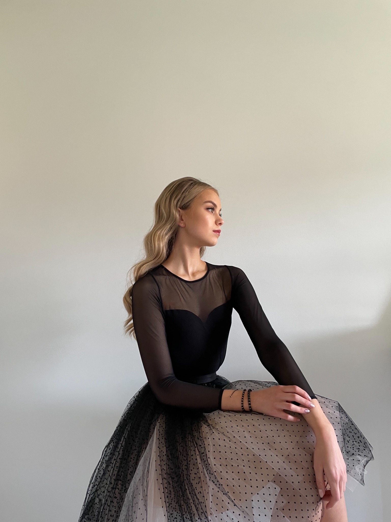 Portret kobiety w czarnej sukience z tiulowym dołem w kropki, siedzącej na tle jasnej ściany i patrzącej w bok, z delikatnym makijażem i blond, falowanymi włosami.