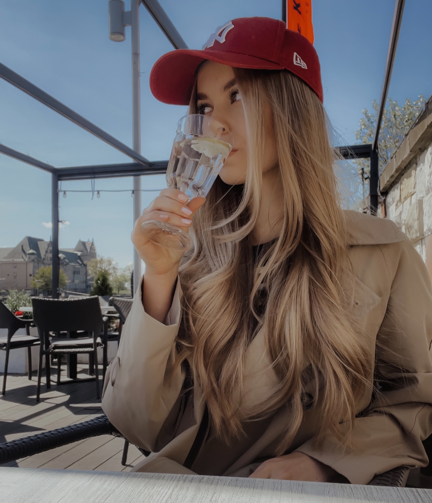 Blondwłosa kobieta w beżowym płaszczu i czerwonej czapce pije wodę z cytryną na tarasie kawiarni z widokiem na miasto.