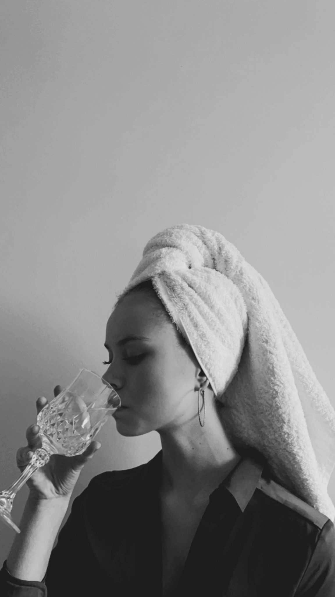 Czarno-białe zdjęcie kobiety z ręcznikiem na głowie, pijącej wodę z kryształowego kieliszka, z widocznym kolczykiem w kształcie koła.