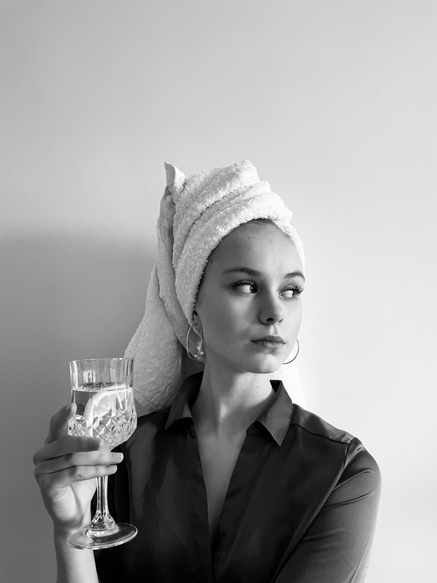 Czarno-białe zdjęcie kobiety z ręcznikiem na głowie, trzymającej kryształowy kieliszek z wodą i cytryną, ubrana w satynową koszulę, z kolczykami w kształcie kół, spoglądającej w bok.