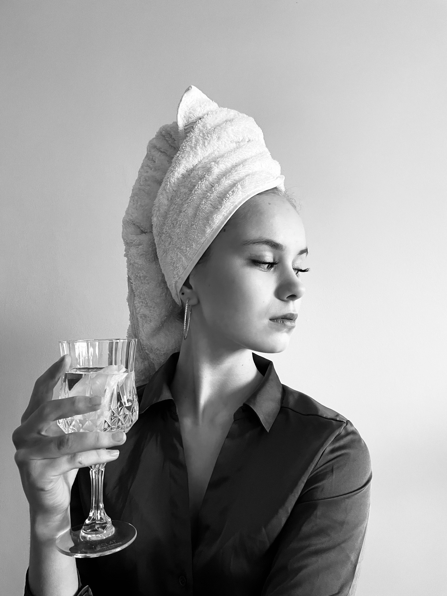 Czarno-białe zdjęcie kobiety z ręcznikiem na głowie, trzymającej kryształowy kieliszek z wodą, patrzącej w bok.