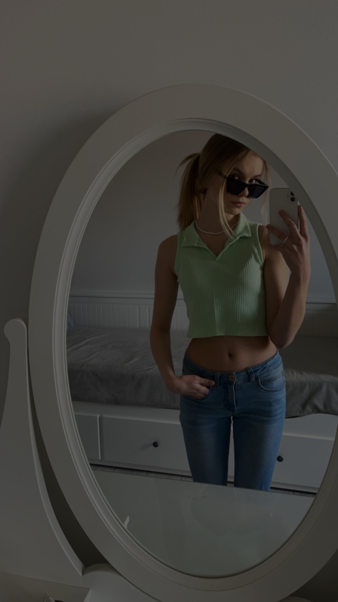 Młoda kobieta w okularach przeciwsłonecznych robi selfie w lustrze w sypialni, ubrana w zielony top i jeansy.