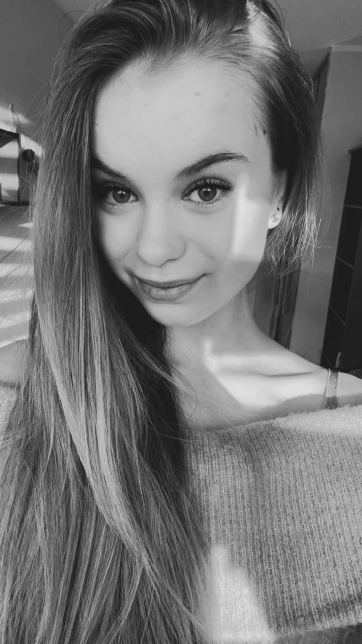 Portret młodej kobiety z długimi włosami, uśmiechniętej, w swetrze z odkrytymi ramionami, w czerni i bieli z naturalnym oświetleniem.