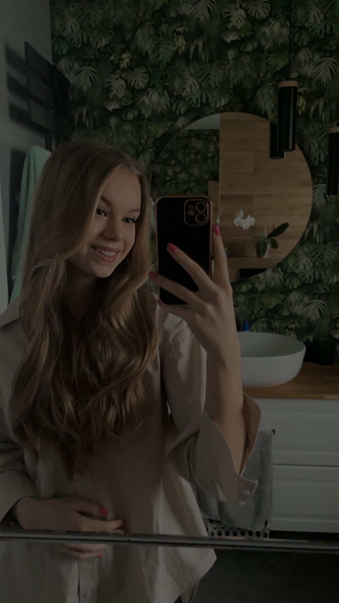 Uśmiechnięta młoda kobieta robi selfie w łazience z tropikalną tapetą, okrągłym lustrem i białą umywalką.