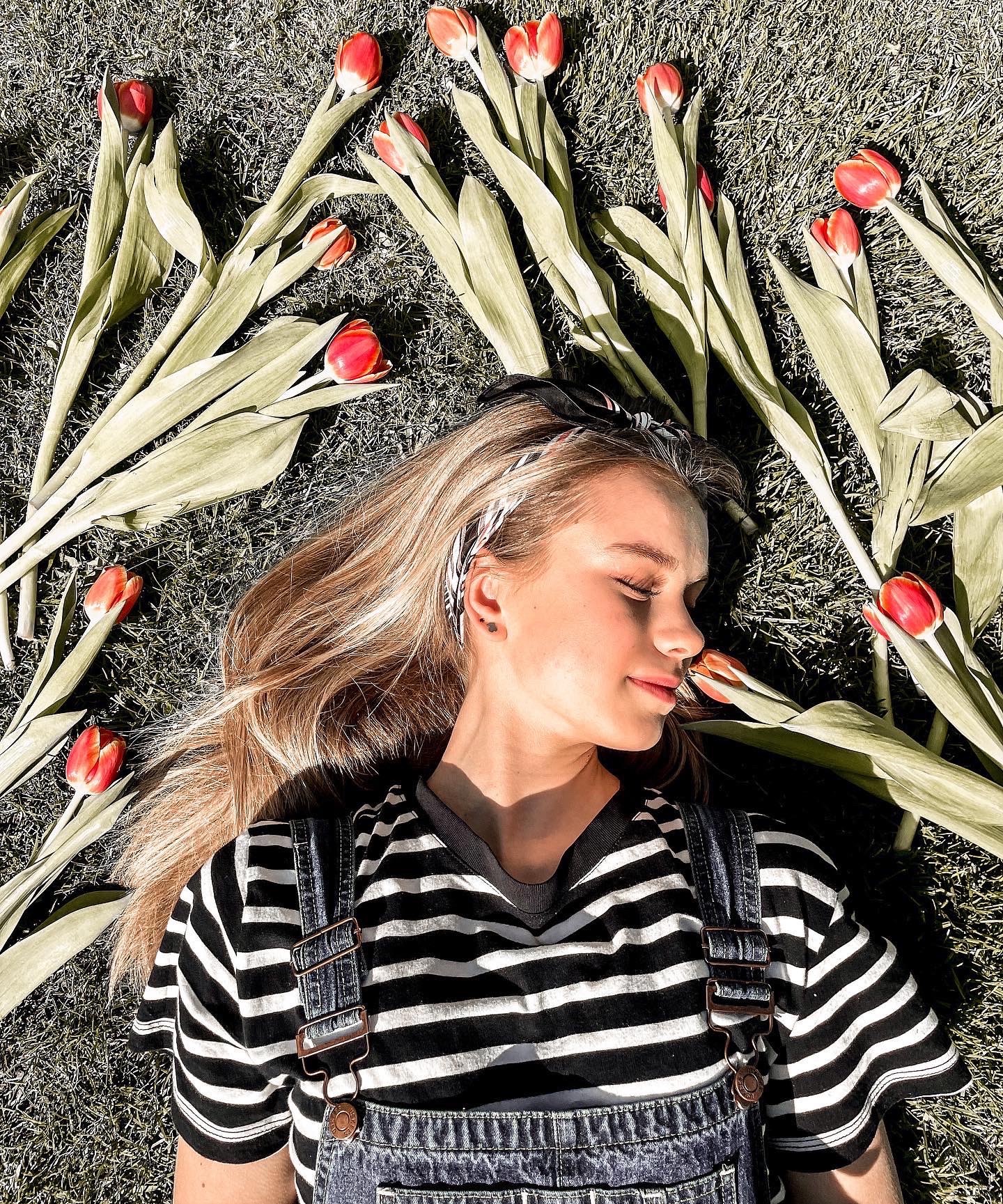 Młoda kobieta w dżinsowych ogrodniczkach i bluzce w paski leży na trawie otoczona czerwonymi tulipanami, z zamkniętymi oczami i delikatnym uśmiechem.