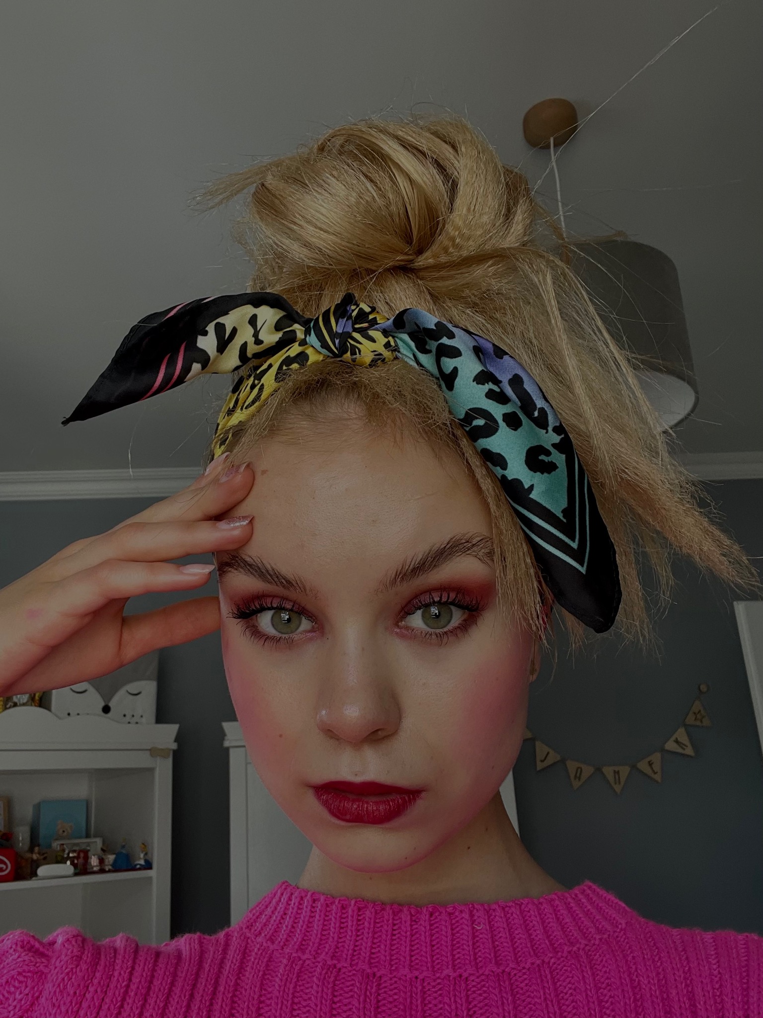 Portret młodej kobiety z blond włosami upiętymi w kok, w różowym swetrze i z kolorową apaszką na głowie. Wyrazisty makijaż oczu i ust. Ujęcie z bliska.