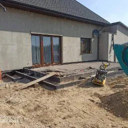 Budowa tarasu z kostki brukowej przy nowym domu: przygotowane podłoże, obrzeża, ubijarka i ramię koparki w kadrze.