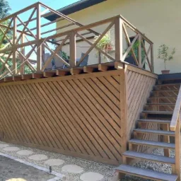 Drewniana altana z ażurową ścianą i schodami, prowadząca na taras z roślinami doniczkowymi, widok z poziomu ogrodu z kamienną ścieżką.