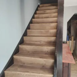 Schody wyłożone panelami drewnopodobnymi z ciemną listwą przy ścianie i ciemną balustradą, widok z dołu.