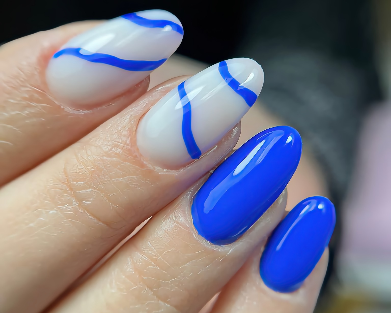 Zbliżenie na dłoń z manicure hybrydowym: dwa paznokcie w kolorze intensywnego błękitu, dwa w kolorze białym z błękitnymi, asymetrycznymi paskami.