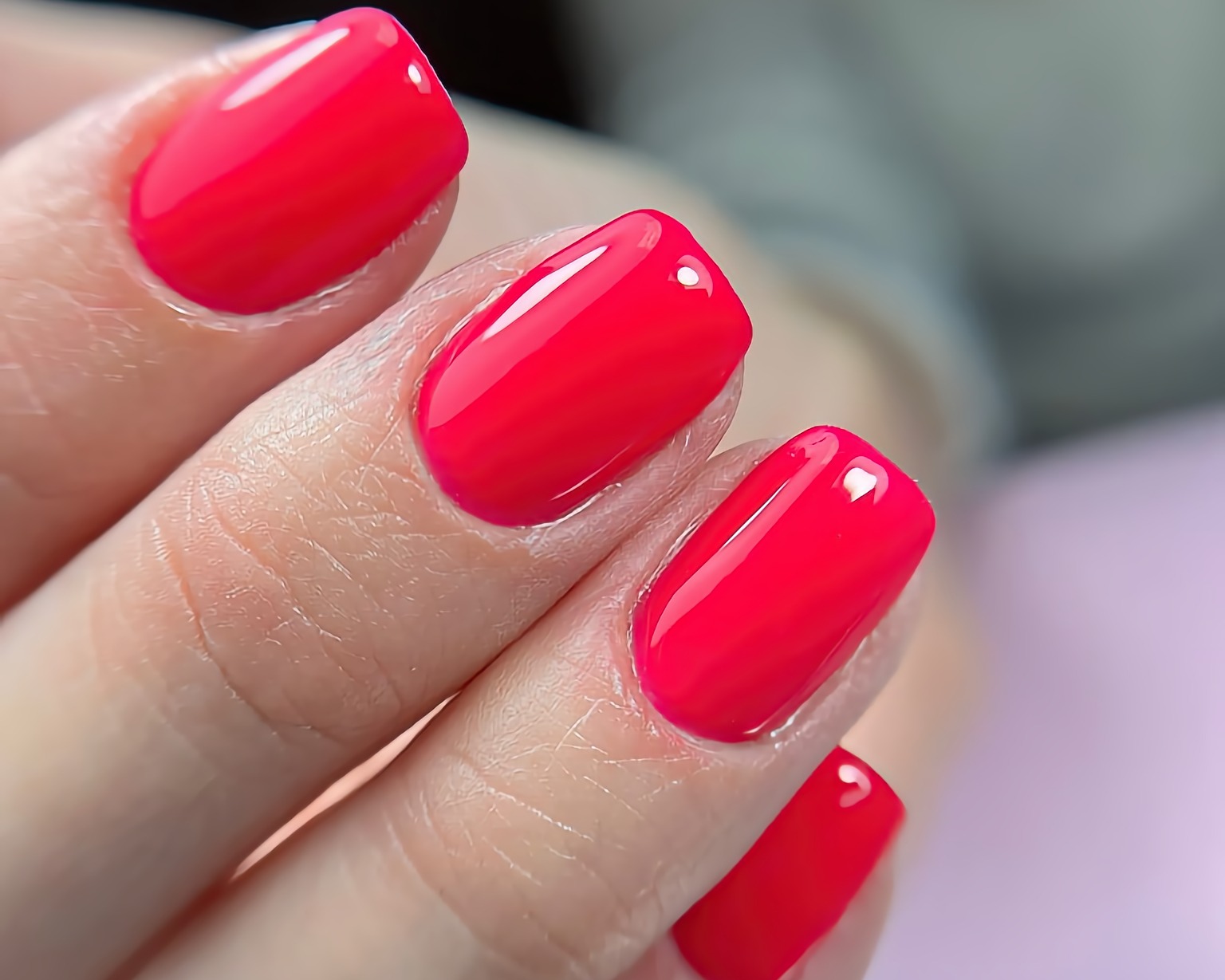 Zbliżenie na dłoń z jaskrawoczerwonym, błyszczącym manicurem hybrydowym o kwadratowym kształcie, widoczne delikatne skórki i refleksy światła na paznokciach.