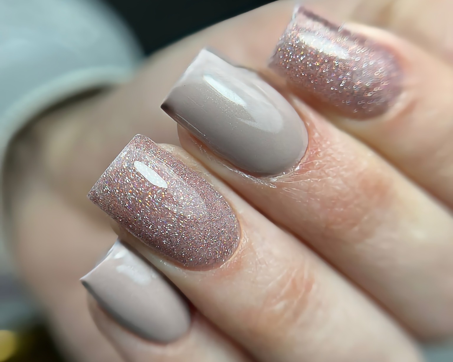 Powiększenie dłoni z kwadratowymi paznokciami: dwa w kolorze taupe, dwa pokryte brokatowym lakierem w odcieniu różowego złota.