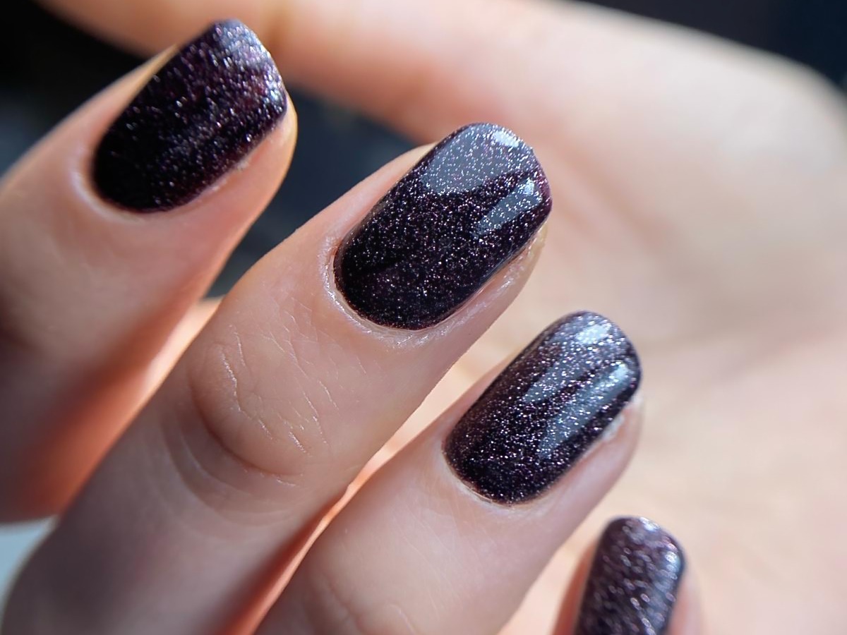 Zbliżenie dłoni z ciemnym, brokatowym lakierem hybrydowym na kwadratowych paznokciach.