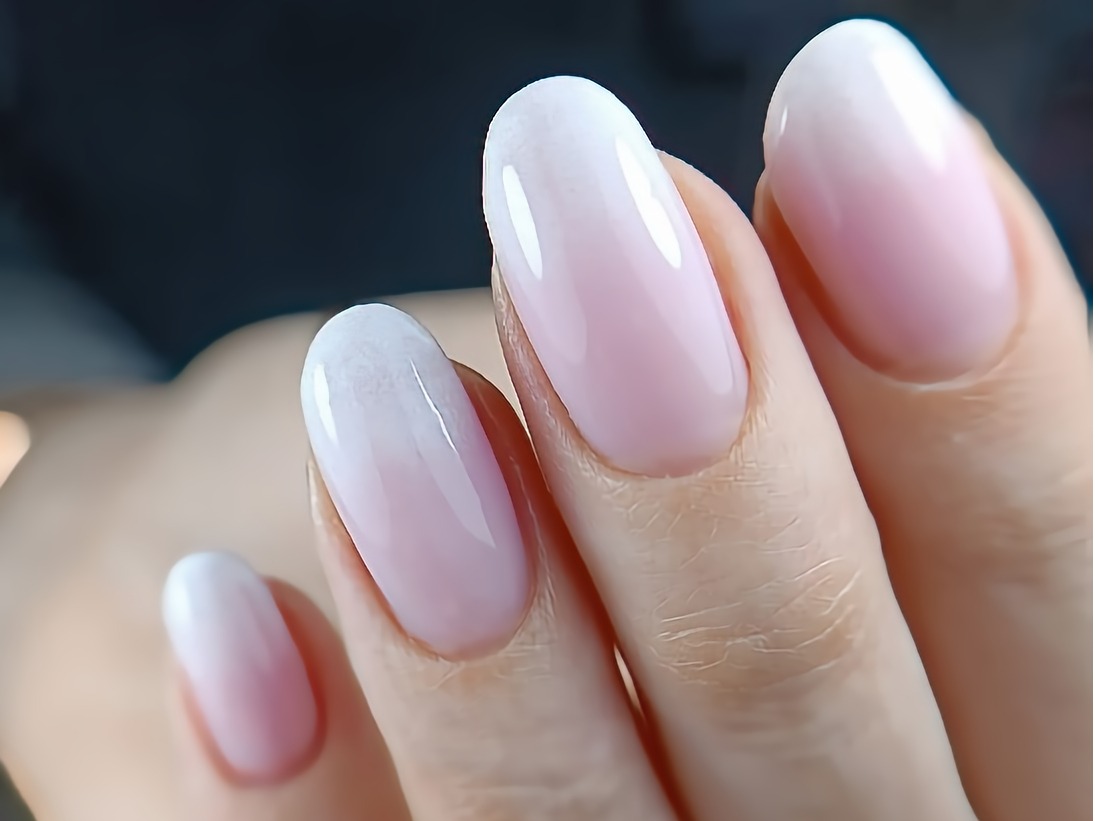 Delikatne ombre na owalnych paznokciach, przechodzące od bieli na końcach do jasnego różu u nasady, na tle ciemnego, rozmytego tła.