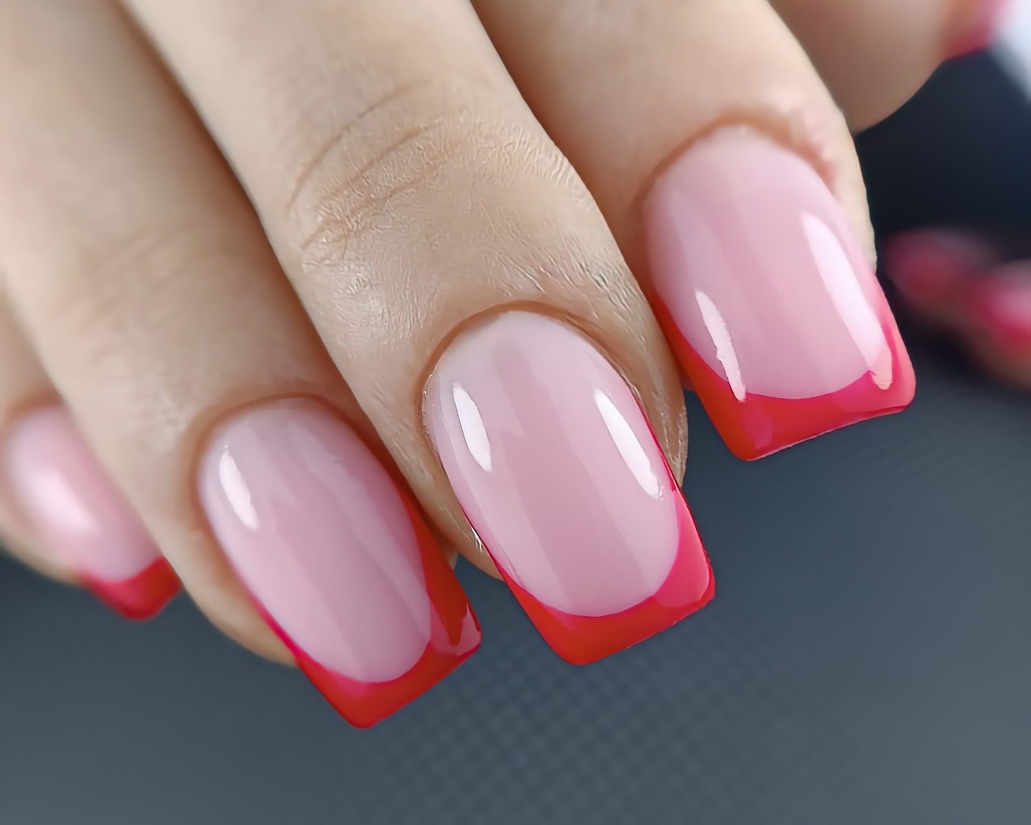 Powiększone ujęcie dłoni z pomalowanymi na jasny róż i czerwony kolor paznokciami w stylu french manicure, na szarym tle.