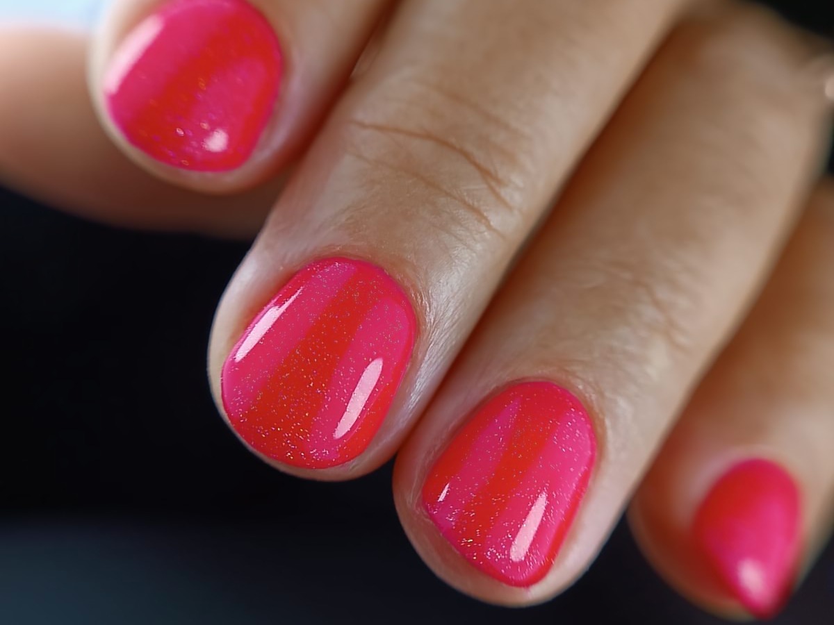 Zbliżenie dłoni z pomalowanymi na intensywny, błyszczący róż paznokciami, z delikatnym efektem ombre przechodzącym w odcień pomarańczowy przy skórkach.