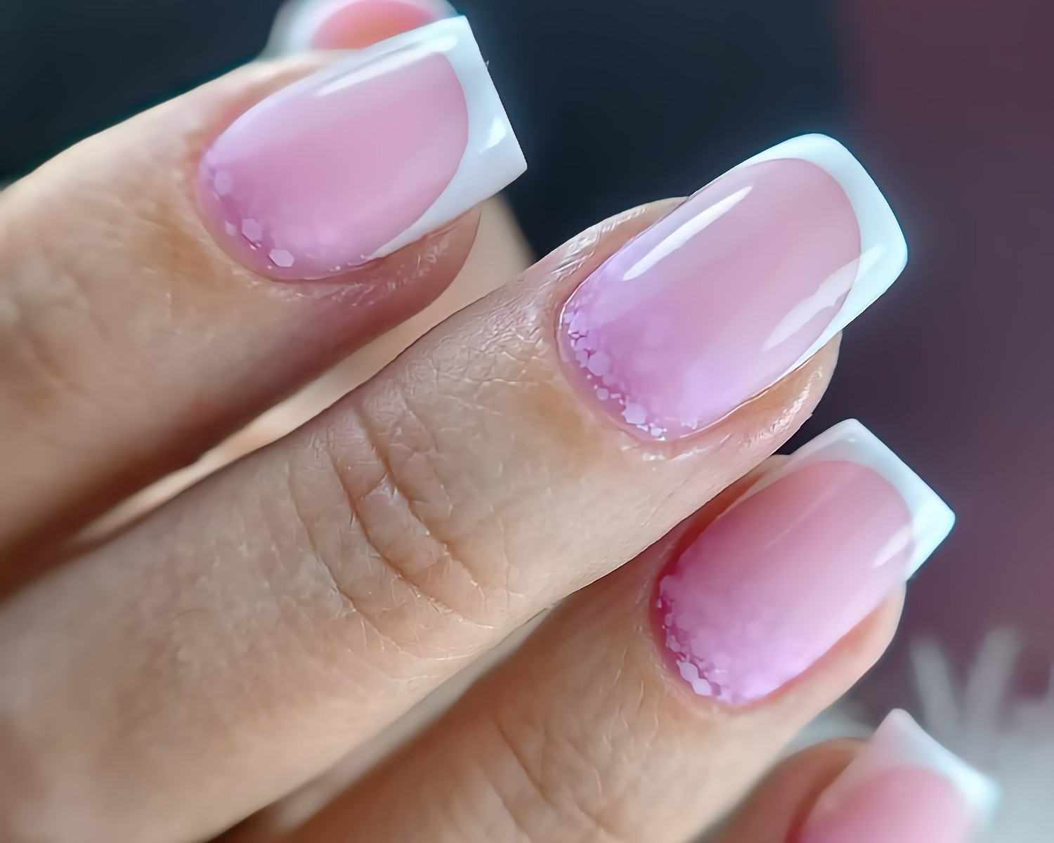 Zbliżenie dłoni z kwadratowymi paznokciami w stylu french manicure, z delikatnym różowym gradientem i drobnymi białymi zdobieniami przy skórkach, na rozmytym tle.