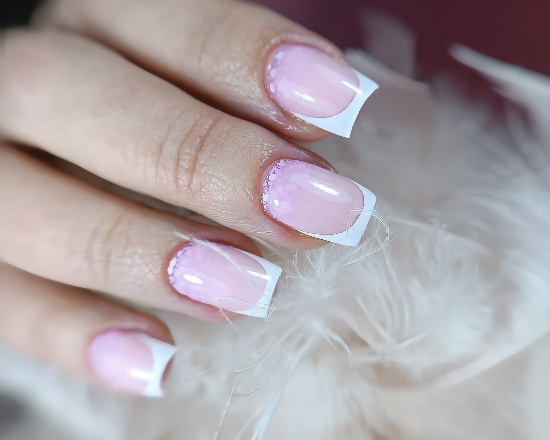 Delikatny manicure francuski z białymi końcówkami i subtelnym, brokatowym zdobieniem przy skórkach, na tle białych piór.