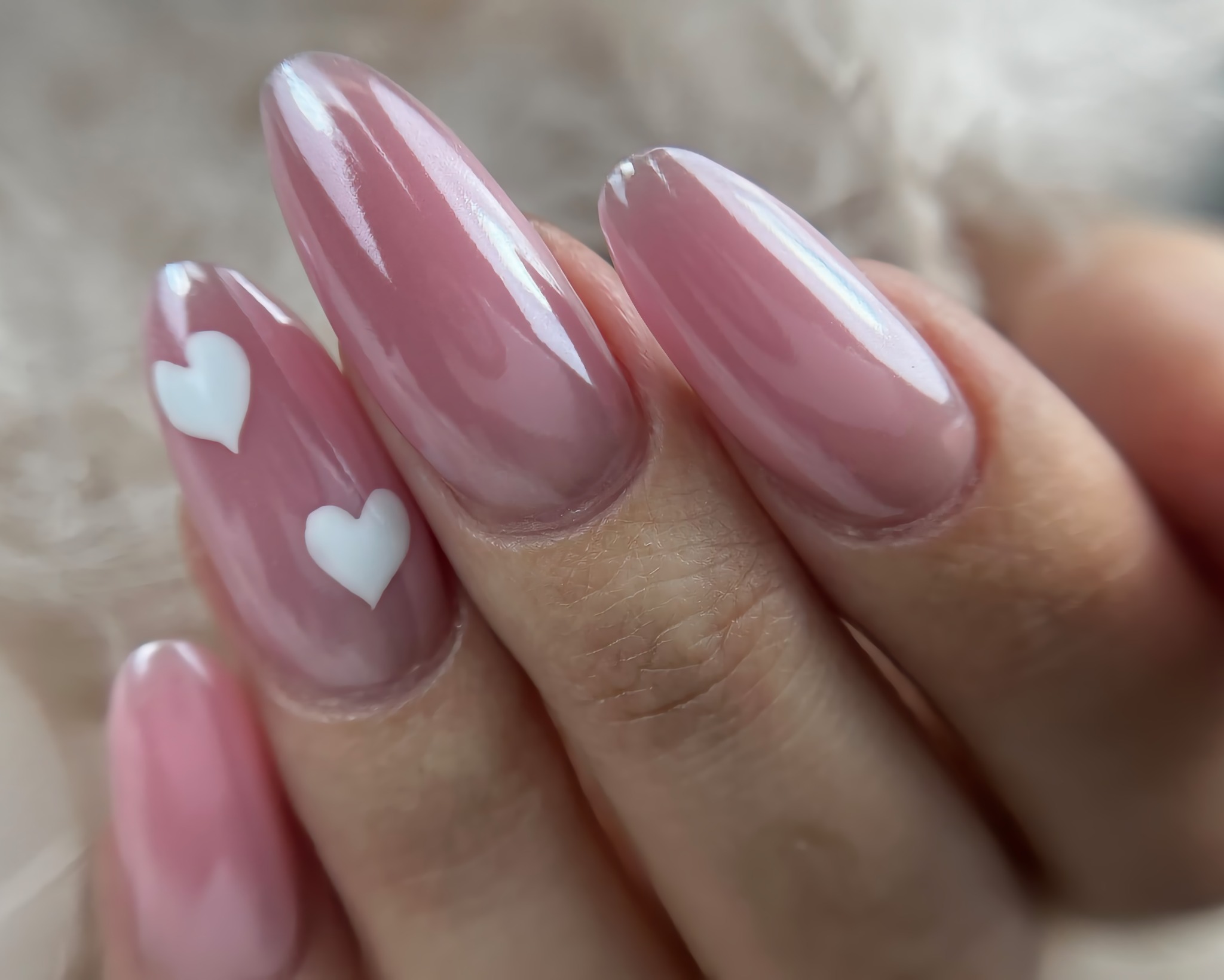 Delikatne, różowe paznokcie w kształcie migdała z białymi serduszkami na tle puszystej tkaniny.