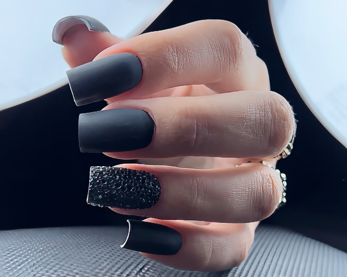 Zbliżenie dłoni z pomalowanymi na czarno paznokciami o kwadratowym kształcie, jeden z paznokci zdobiony czarnymi koralikami, matowe wykończenie.