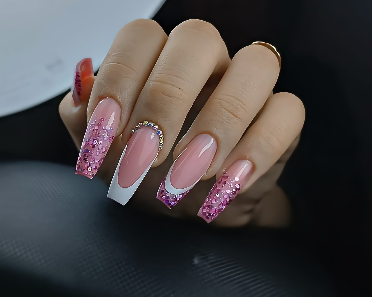 Dłoń z długimi paznokciami: dwa z brokatem w odcieniach różu, jeden w stylu french manicure z brokatową końcówką, a jeden ozdobiony cyrkoniami.