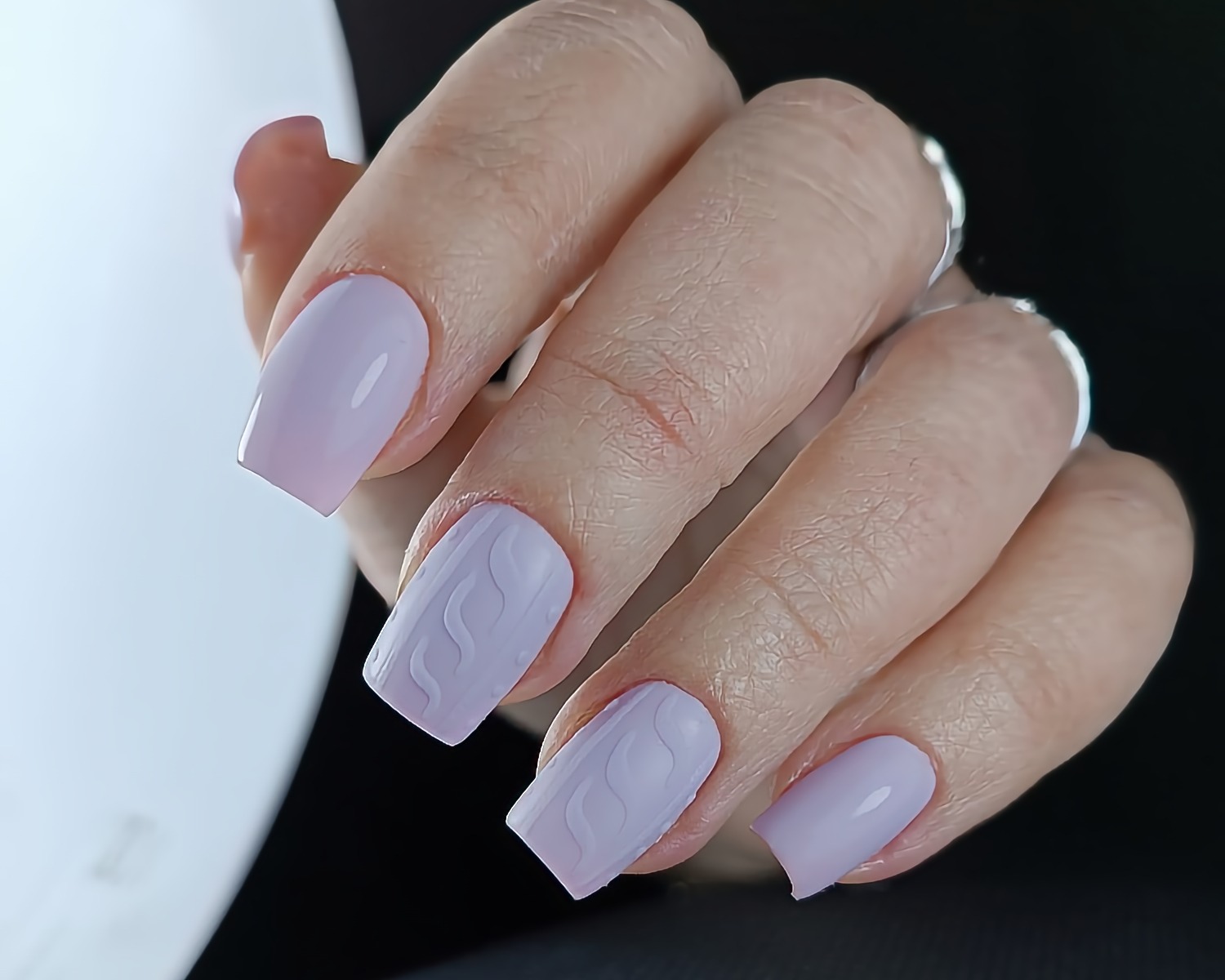 Dłoń z pomalowanymi na jasnofioletowo paznokciami, dwa z nich z wzorem imitującym splot swetra.