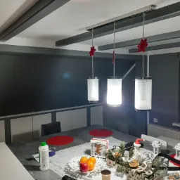 Szare wnętrze z pomalowanymi na szaro ścianami, sufitem z dekoracyjnymi belkami i trzema wiszącymi lampami z czerwonymi ozdobami, stół z dekoracjami świątecznymi i butelką z zieloną nakrętką.