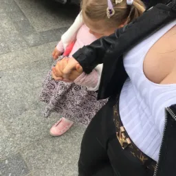 Dziewczynka z blond włosami spiętymi w dwa kucyki, trzymająca za rękę dorosłą osobę na chodniku z szarych płyt.