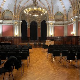 KORSAR Grzegorz Korczyński - Sala koncertowa z parkietem w jodełkę, przygotowana na wydarzenie, z rzędami czarnych krzeseł i kartonami w tle, oświetlona ozdobnym żyrandolem.