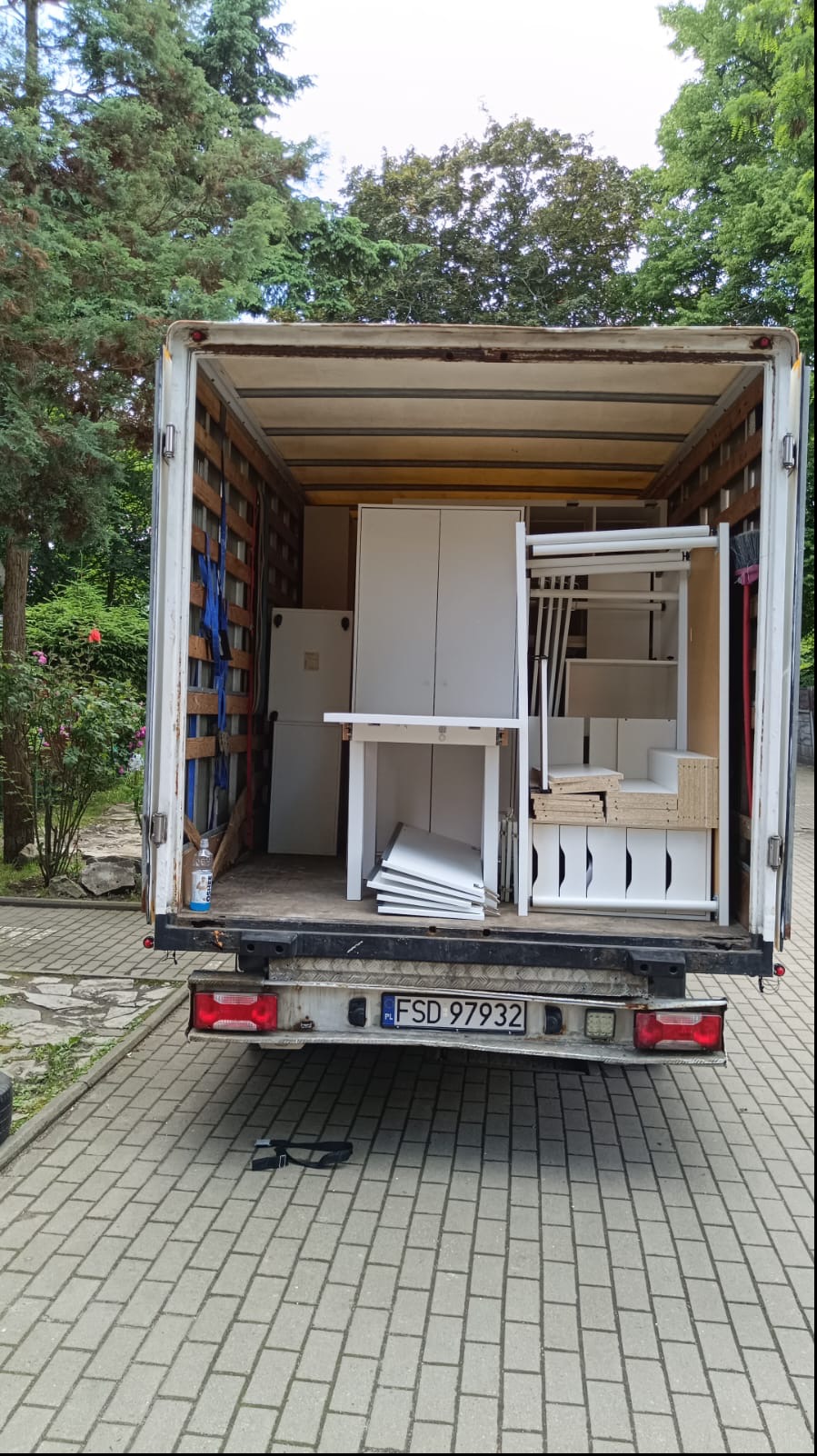 Otwarta przestrzeń ładunkowa ciężarówki wypełniona białymi meblami, w tym szafą, stołem i regałami, widoczne tablice rejestracyjne pojazdu na tle brukowanej powierzchni.