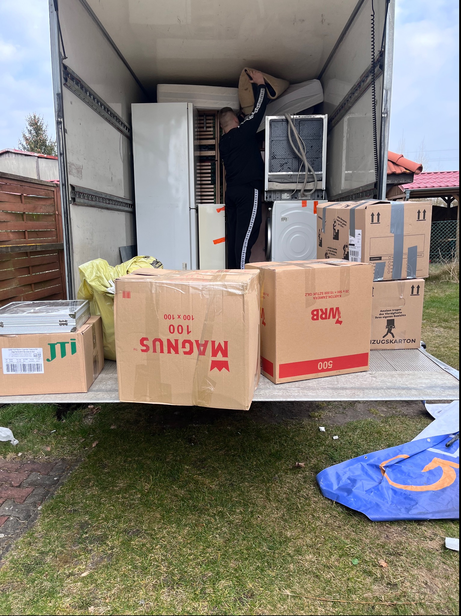 Załadunek mebli i sprzętów AGD do wnętrza samochodu dostawczego, widoczne kartony z napisami Magnus i WRB, osoba w czarnym dresie układa przedmioty wewnątrz