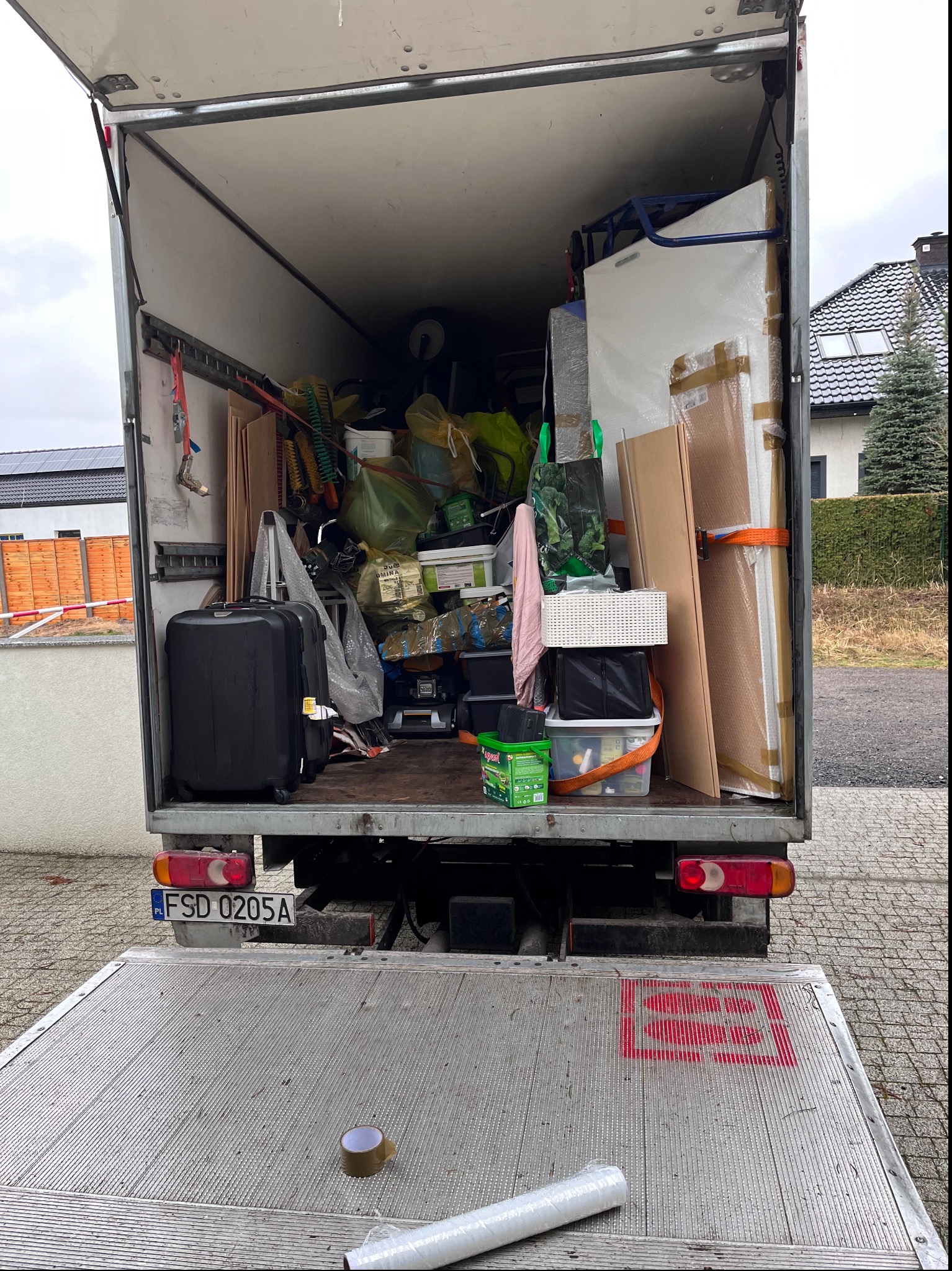 Wnętrze załadowanego busa transportowego z widocznymi kartonami, meblami, walizkami i różnymi przedmiotami zabezpieczonymi taśmami, widok od tyłu z otwartą platformą załadunkową i fragmentem ulicy...