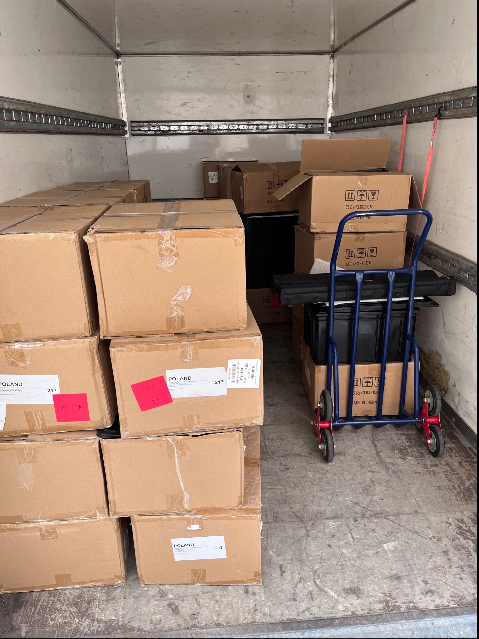 Wnętrze furgonetki transportowej wypełnionej kartonami z naklejkami 'Poland' i 'Made in China', część kartonów zabezpieczona taśmą, obok wózek transportowy z czarnymi przedmiotami, widoczne ślady...
