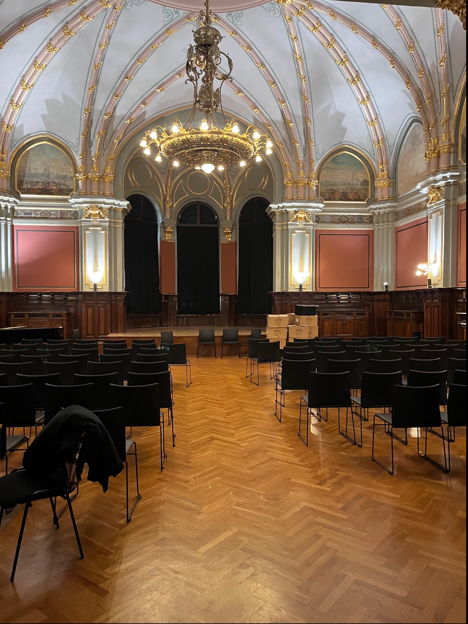 Sala koncertowa z parkietem w jodełkę, przygotowana na wydarzenie, z rzędami czarnych krzeseł i kartonami w tle, oświetlona ozdobnym żyrandolem.