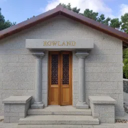 Małe kamienne mauzoleum z drewnianymi drzwiami zdobionymi geometrycznym wzorem, zwieńczone napisem 'ROWLAND' w złotych literach, otoczone zielenią drzew i trawnika.